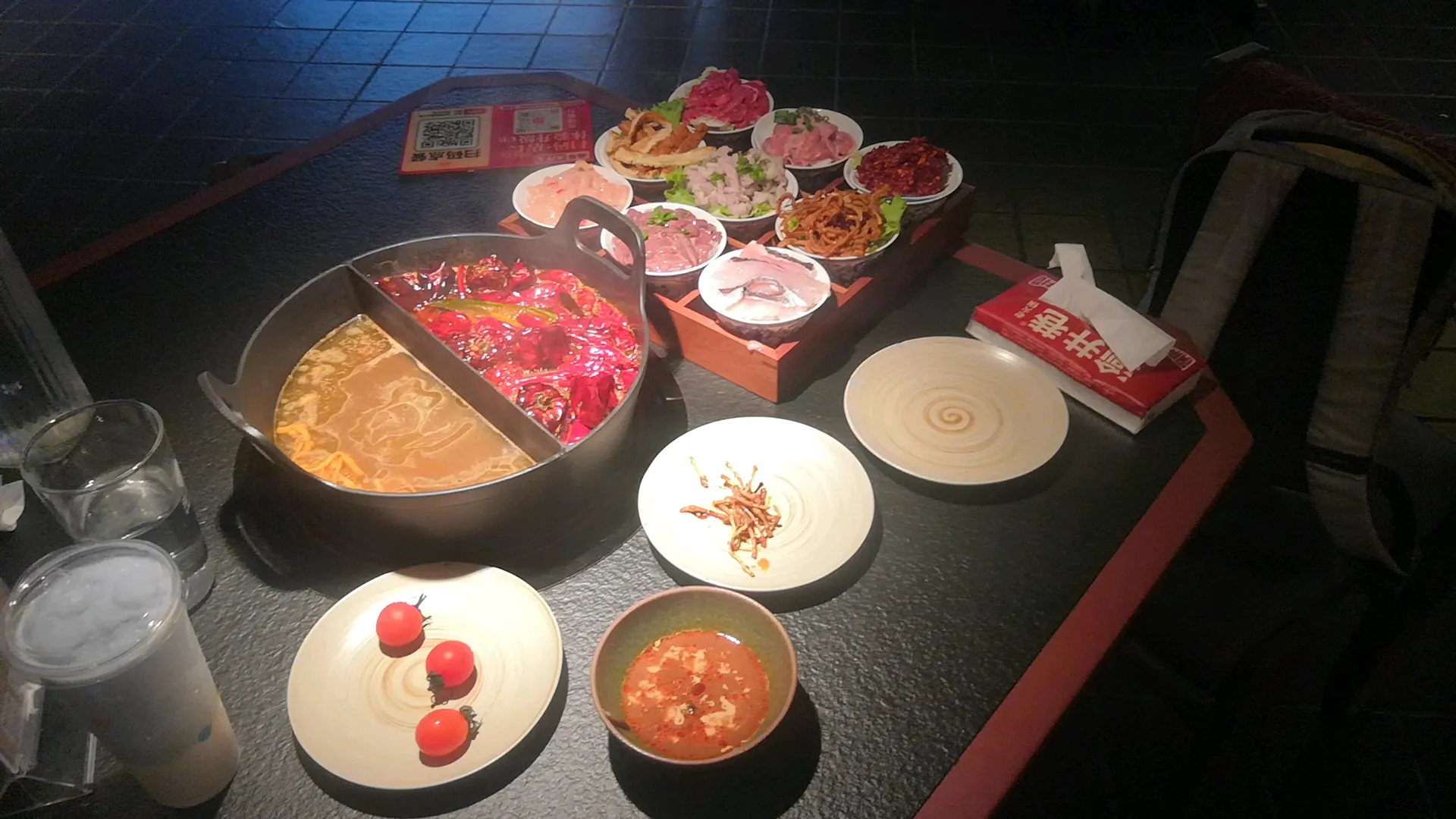 A divided hotpot with red and mild broths, ingredients, and dipping sauces on a dining table.