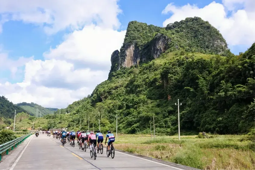 A group of cyclists riding along a road beside green limestone hills.